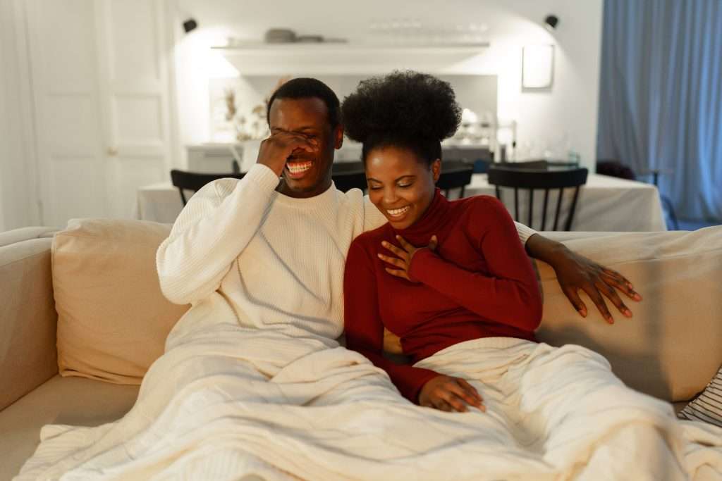 The Love Central - Happy young african american couple laughing at funny joke watching comedy movie together at home The Love Central -