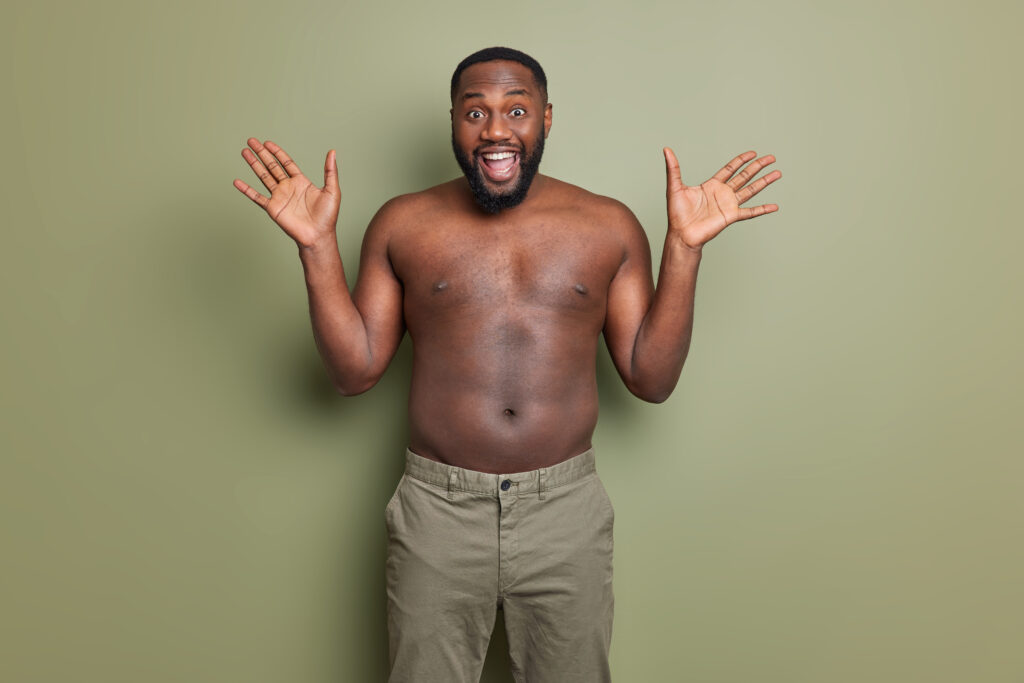 The Love Central - Happy emotional man with dark skin raises palms reacts happily on unexpected surprise smiles broadly stands shirtless indoor gets nice offer opens mouth in excitement enjoys big sales in shop Body image is how you see yourself when you look in the mirror