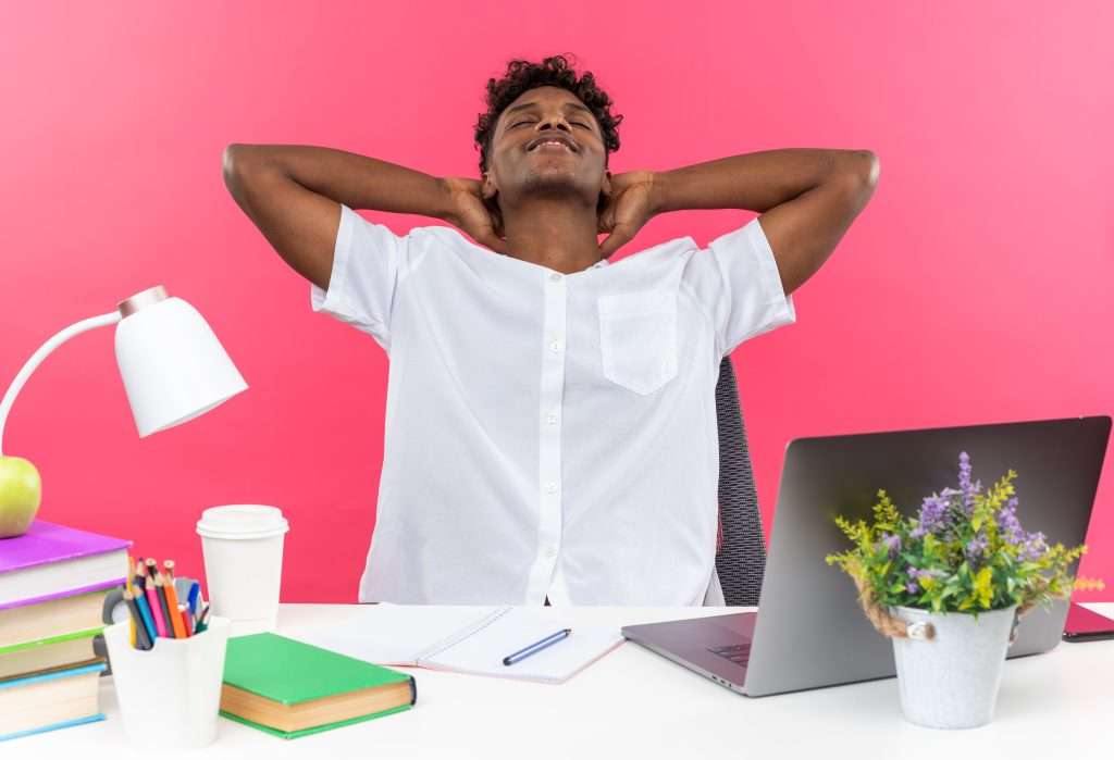 The Love Central - pleased young afro-american student sitting with closed eyes at desk with school tools putting hands on his head isolated on pink background The Love Central -