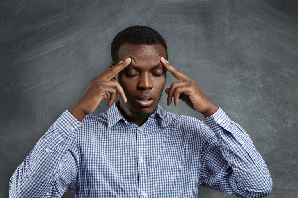 The Love Central - Handsome serious puzzled African student dressed in checkered shirt robbing his forehead closing his eyes looking concentrated and focused trying to remember right answer during test in classroom The Love Central -