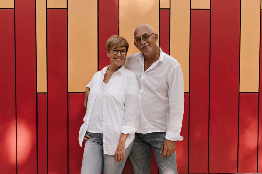 The Love Central - Stylish woman with short hair in eyeglsses and light blouse smiling and hugging with old man in white shirt and jeans on red and orange background The Love Central -
