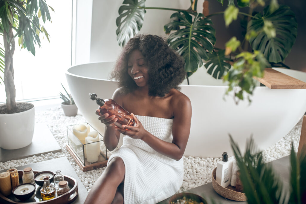 The Love Central - Pretty dark-skinned woman civered with towel choosing body lotion The Love Central -