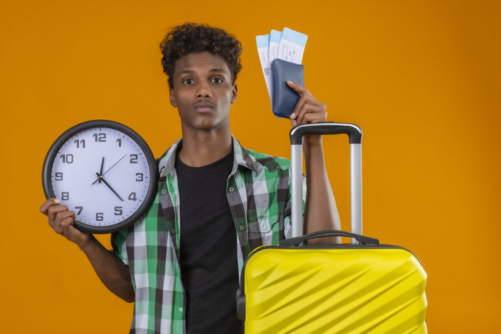 The Love Central - young african american traveler man standing with suitcase holding air tickets and clock looking worried and confused over orange background The Love Central -