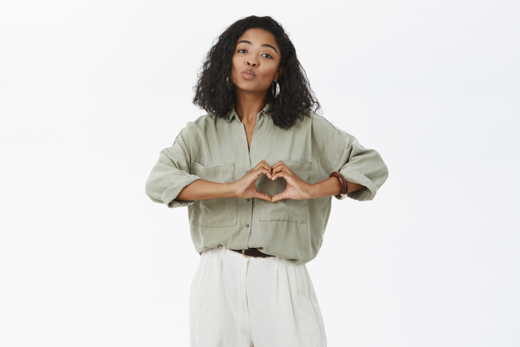 The Love Central - Woman wants share love with close people. Tender attractive african american female in trendy shirt and pants folding lips in mwah gesture showing heart sign over chest expressing affection The Love Central -