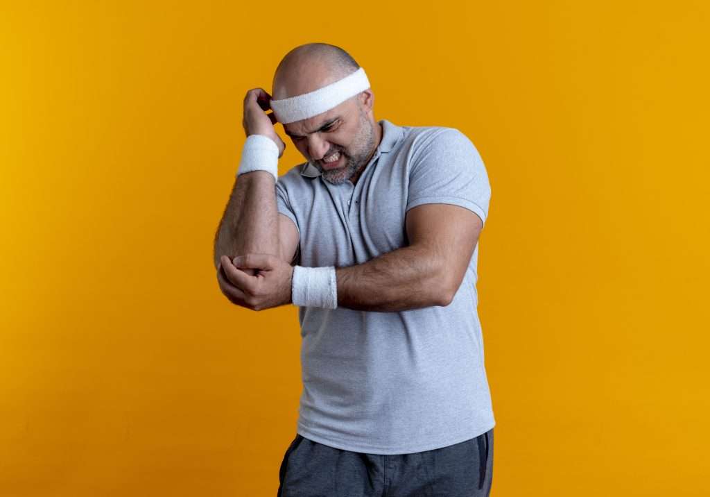 The Love Central - mature sporty man in headband touching elbow looking unwell feeling pain standing over orange background The Love Central -