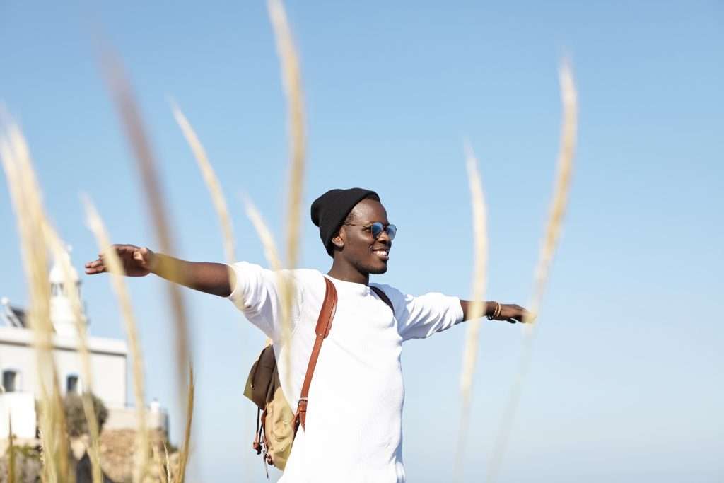 The Love Central - Youth, happiness, freedom and traveling. Cheerful young African American man traveler wearing hat and sunglasses spreading arms and smiling happily, feeling relaxed and in harmony with nature The Love Central -