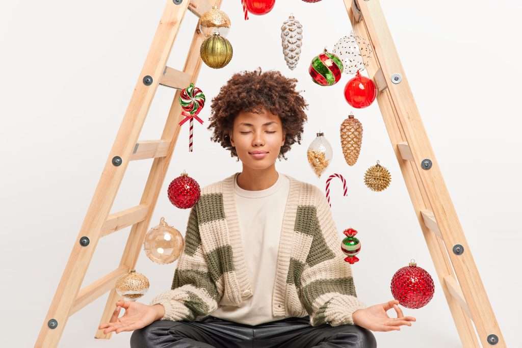 The Love Central - Dark skinned woman sits in lotus pose keeps eyes closed breathes deeply has meditation exercise tries to relax in tranquil atmosphere poses against New Year background practices yoga at home Essential List of Healthy Habits to Beat Holiday Stress.