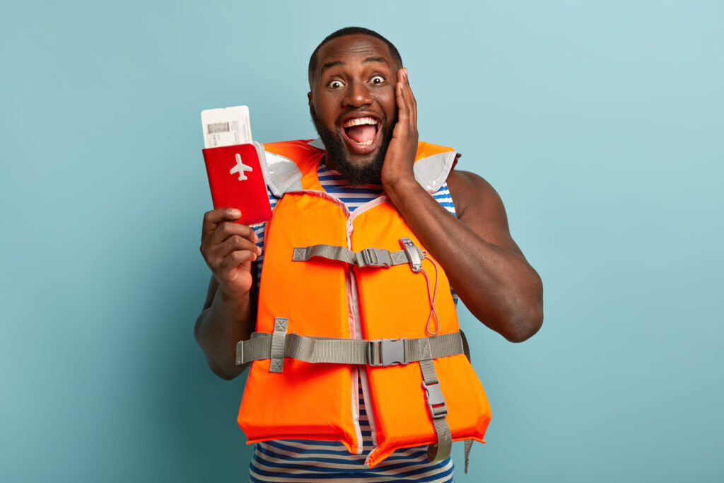 The Love Central - Half length shot of happy black man enjoys coming travel, holds red passport with tickets, shares impressions, wears lifejacket, touches cheek, isolated on blue studio wall. Summer time and tourism The Love Central -