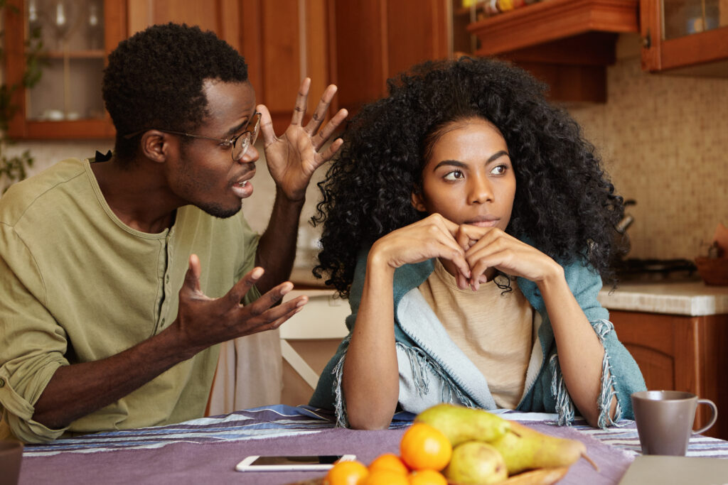 The Love Central - People and relationships concept. African American couple arguing in kitchen: man in glasses gesturing in anger and despair, screaming at his beautiful unhappy girlfriend who is totally ignoring him Financial Disparity in Relationships