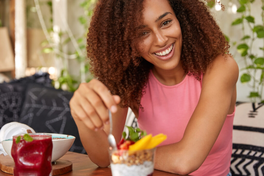 The Love Central - Happy African woman with curly hair, eats delicious dessert, holds spoon, dressed in casual pink vest, enjoys pleasant conversation with friend, poses against cozy interior. Time for lunch and rest If you're someone who feels like they're constantly running at high speed, with a mind that's always racing, you might have an excess of vata dosha