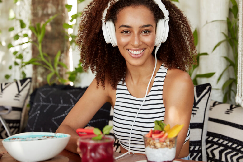 The Love Central - Beautiful positive African American woman enjoys listening music in headphones, spends leisure time at loft cafeteria with cocktail, has break after work, demonstrates pleasant smile. The Matterhorn Method