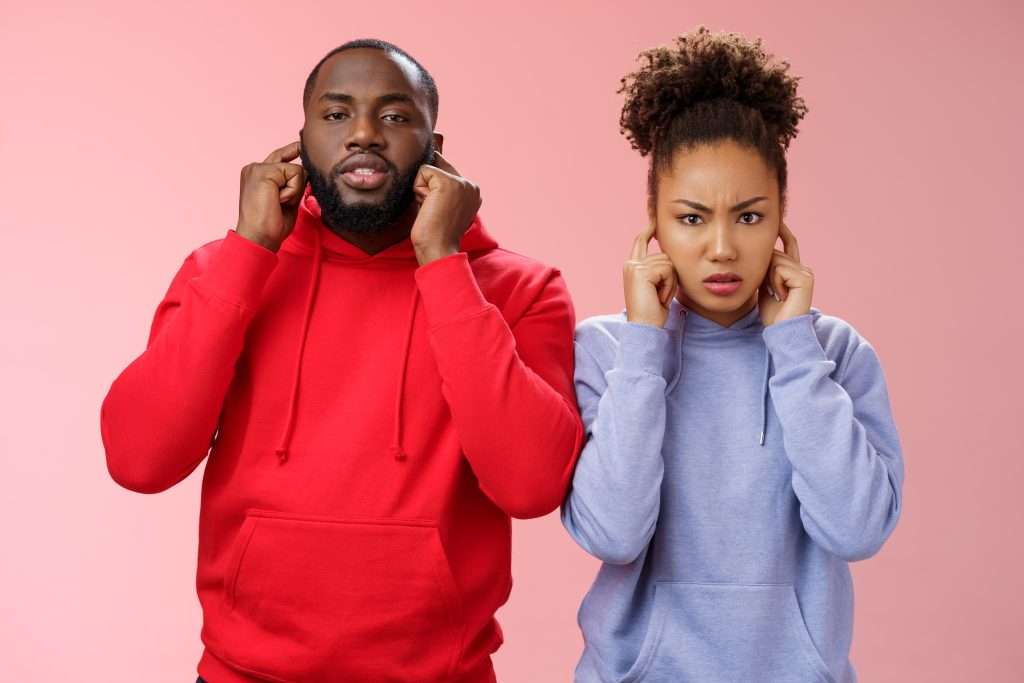The Love Central - Annoyed tired couple african american woman man relationship exhausted each other close ears plug index fingers frowning look irritated hear terrible disturbing noise, standing pink background The Love Central -