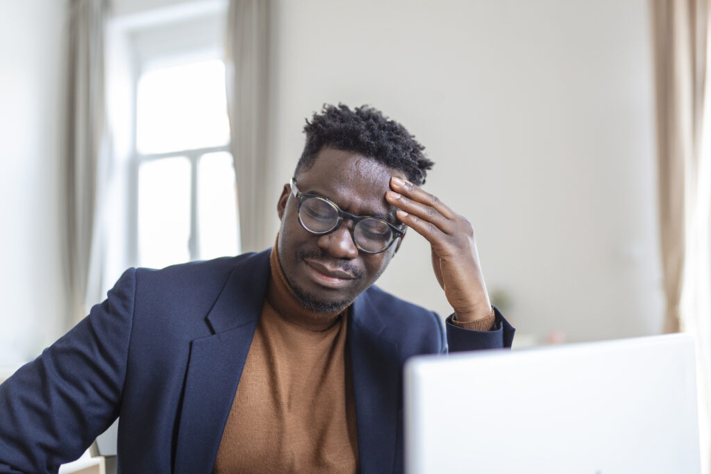 The Love Central - Head shot stressed young african american man touching forehead, suffering from terrible headache, working on computer at home office. Frustrated confused biracial guy having painful feelings in head. A