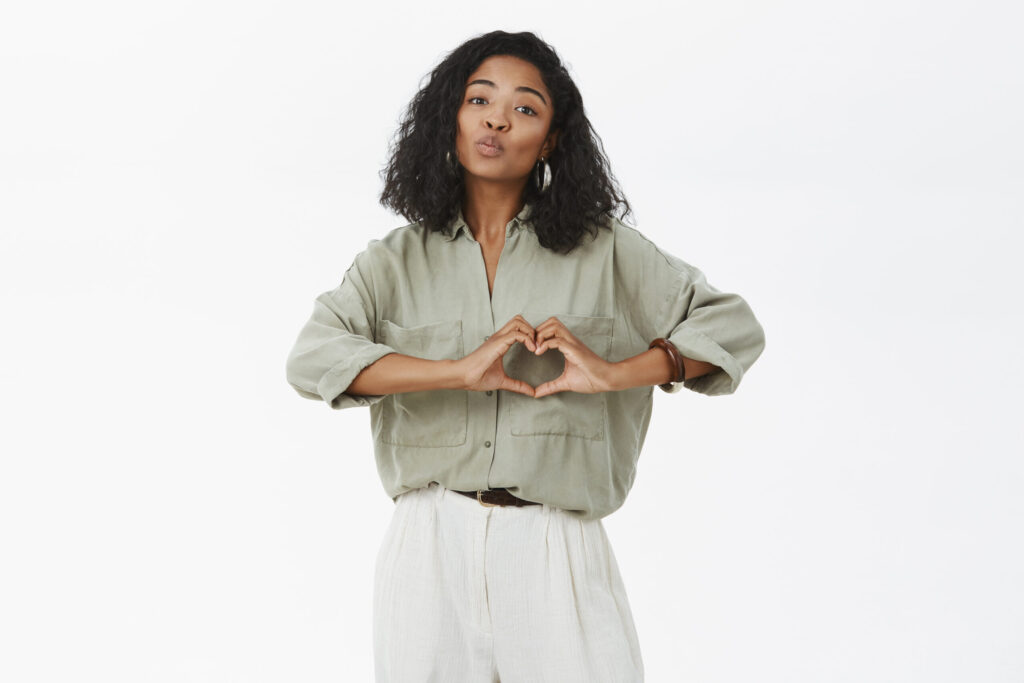 The Love Central - Woman wants share love with close people. Tender attractive african american female in trendy shirt and pants folding lips in mwah gesture showing heart sign over chest expressing affection The Love Central -