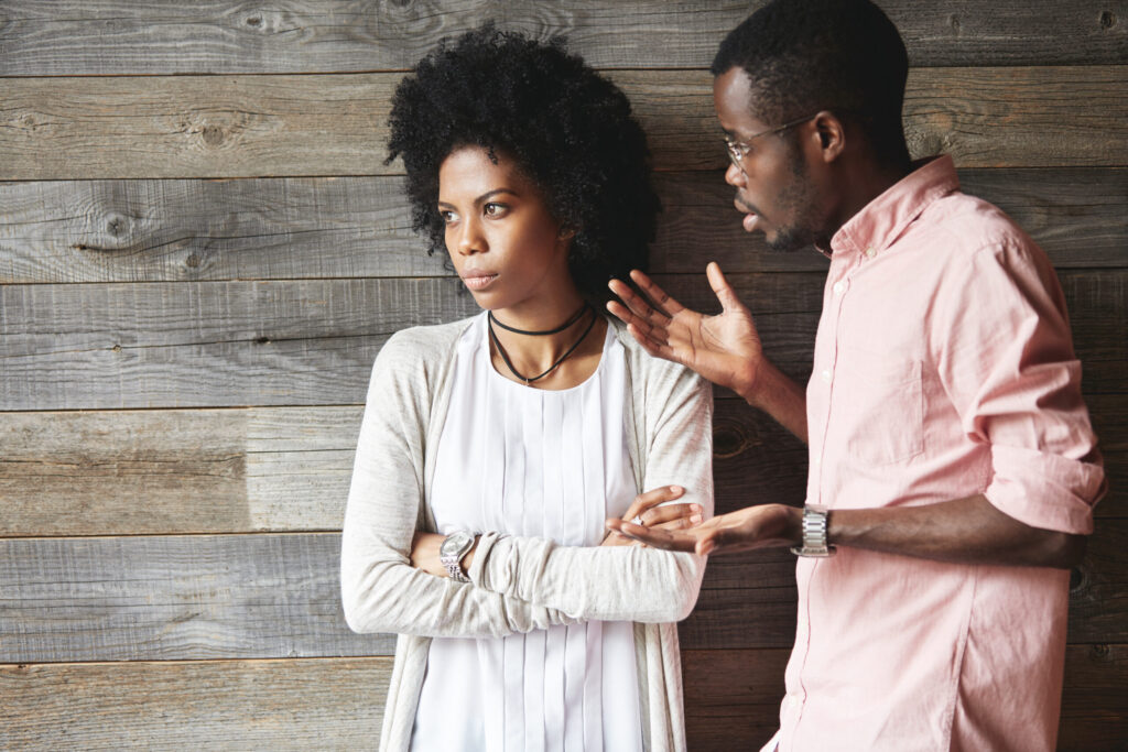 The Love Central - Young displeased black male gesturing in indignation, arguing with his stylish girlfriend, who is standing against wooden wall with folded arms, looking away with offended expression on her face The Love Central -