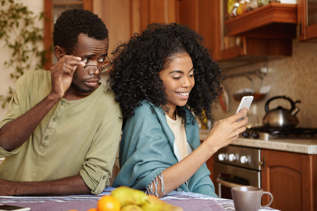 The Love Central - Jealous curious black man holding glasses spying his girlfriends mobile phone while she is typing message to her lover and smiling happily Betrayal unfaithfulness infidelity and lack of trust The Love Central -